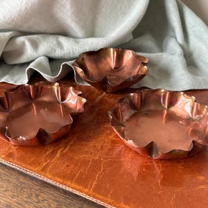 May include: Three small, copper-colored bowls with scalloped edges. The bowls are arranged on a brown surface, with a light green cloth in the background. The bowls have a shiny, reflective surface.