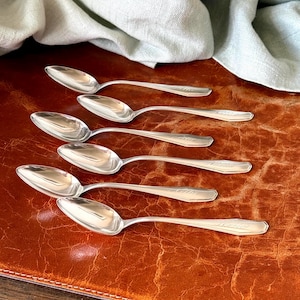 May include: A set of six silver-toned metal spoons arranged in a staggered formation. Each spoon features a smooth, oval-shaped bowl and a slender handle with a subtle, engraved decorative detail near the end of the handle stem.