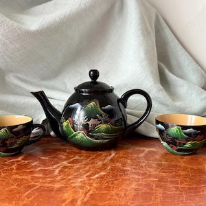 May include: A black tea set featuring a teapot and two cups. The set is decorated with a landscape scene of mountains, trees, and buildings in shades of green, red, and gold. The cups and teapot have a capacity of approximately 6-8 ounces.