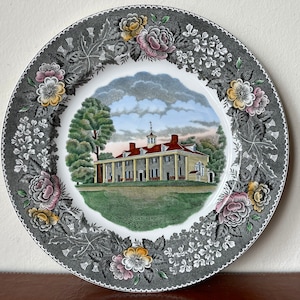 May include: A gray ceramic plate with a floral border and a central image of a white house with a red roof. The house is surrounded by green trees and grass. The text "Mount Vernon" is printed below the image.