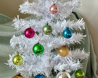 Pode incluir: Uma pequena árvore de Natal artificial branca decorada com uma variedade de enfeites de vidro coloridos. Os enfeites são em tons de vermelho, rosa, verde, dourado e azul. A árvore está sobre um fundo verde suave.