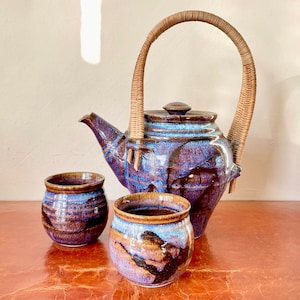 May include: A brown and blue ceramic teapot with a woven handle and two matching ceramic cups. The teapot and cups have a speckled glaze.