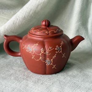 May include: A small, reddish-brown ceramic teapot with a lid. The teapot features a rounded body, a handle, and a spout. Delicate floral designs in gold and blue adorn the surface. The lid has a small knob and decorative perforations. A charming piece for tea lovers.