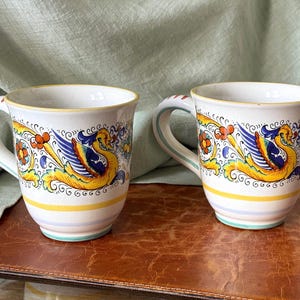 May include: Two ceramic mugs featuring a hand-painted design of a yellow and blue mythical creature with wings, surrounded by orange floral accents and scrolling black lines. The mugs have a white base with yellow and light blue horizontal stripes.