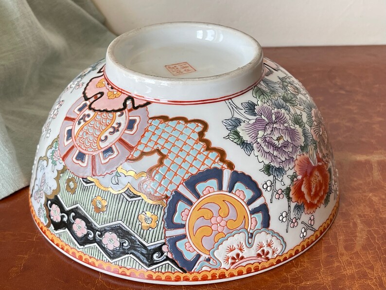 May include: A large white ceramic bowl with a colorful floral and geometric design. The bowl has a wide rim and a rounded bottom. The design features flowers, leaves, and intricate patterns in shades of pink, blue, green, gold, and red.