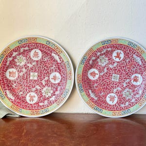 May include: Two round, red and white ceramic plates with intricate floral and geometric patterns. Each plate features Chinese characters and a decorative border with green, yellow, and blue accents. The plates are displayed on a brown surface.
