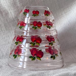 May include: A stack of clear glass bowls with a flared shape. Each bowl is decorated with a pattern of red apples with green leaves. The bowls are nested together, creating a tiered effect.