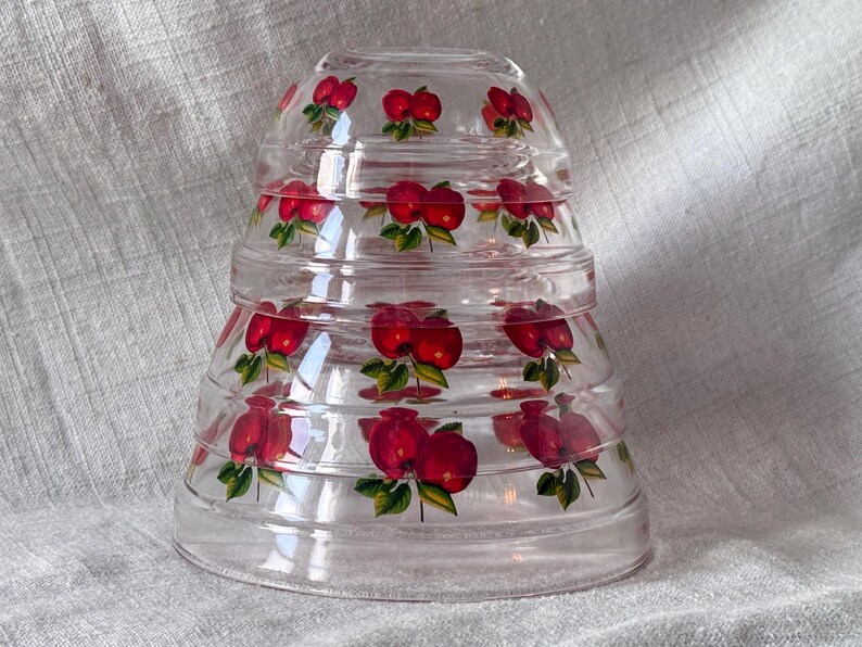 May include: A stack of clear glass bowls with a flared shape. Each bowl is decorated with a pattern of red apples with green leaves. The bowls are nested together, creating a tiered effect.