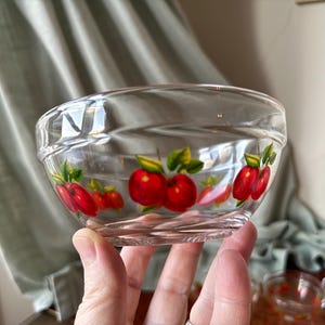 May include: Clear glass bowl with a decorative apple design. The bowl features a series of red apples with green leaves painted around the rim. The bowl is held in a person's hand.