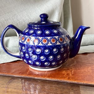 May include: A blue ceramic teapot with a lid, handle, and spout. The teapot is decorated with white and orange floral patterns. It sits on a brown surface with a light green cloth in the background. A classic kitchenware item.