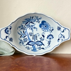 May include: White ceramic serving dish with a blue floral pattern. The dish has a scalloped edge and two handles. The blue floral design features stylized flowers and leaves. The dish is likely used for serving food.
