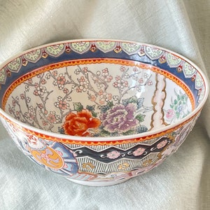 May include: A large, white ceramic bowl with a colorful floral design. The bowl features a red, orange, pink, and blue floral pattern with gold accents. The rim of the bowl is decorated with a blue and gold geometric pattern.