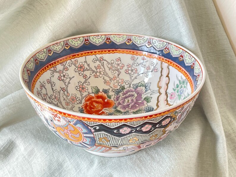 May include: A large, white ceramic bowl with a colorful floral design. The bowl features a red, orange, pink, and blue floral pattern with gold accents. The rim of the bowl is decorated with a blue and gold geometric pattern.