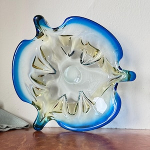 May include: A decorative glass bowl with a unique leaf-like shape. The bowl features a vibrant blue rim that fades into a white and yellow interior. The bowl has a clear, smooth surface and is ideal for displaying small items.
