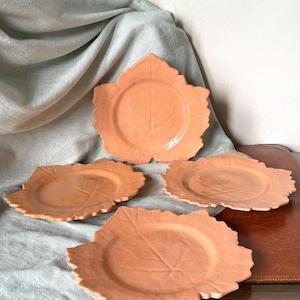 May include: Four peach-colored leaf-shaped plates. Each plate has a central vein design and a scalloped edge. The plates are arranged on a light gray cloth, creating a rustic and natural aesthetic. Ideal for serving salads or appetizers.