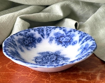 May include: A small, shallow ceramic bowl featuring a white base with a deep blue transferware floral pattern. The design consists of intricate, repeating botanical motifs that cover the interior surface, creating a dense, textured appearance.