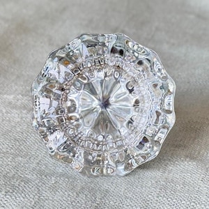 May include: A clear, transparent glass cabinet knob featuring a multi-faceted, starburst-like pattern radiating from the center. The knob has a scalloped, wavy edge and a reflective, polished surface that catches light to create intricate shadows.