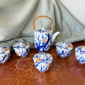 May include: A white porcelain tea set consisting of a teapot with a curved bamboo handle and five matching lidded cups. Each piece features a hand-painted pattern of dark blue branches and small orange and blue flowers across the surface.