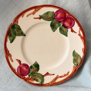 May include: A small, off-white ceramic plate with a decorative border featuring brown branches, green leaves, and red plums. The plate has a simple, classic design, suitable for serving small portions. The plate is approximately 6 inches in diameter.