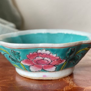 Puede incluir: Un peque&ntilde;o cuenco de cer&aacute;mica blanca con un borde festoneado. El cuenco est&aacute; decorado con un esmalte azul verdoso y un dise&ntilde;o floral pintado a mano en rosa, blanco y verde.