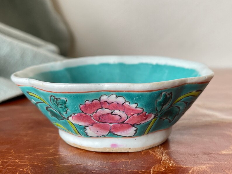 Puede incluir: Un peque&ntilde;o cuenco de cer&aacute;mica blanca con un borde festoneado. El cuenco est&aacute; decorado con un esmalte azul verdoso y un dise&ntilde;o floral pintado a mano en rosa, blanco y verde.
