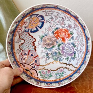 May include: A large white ceramic bowl with a blue and gold rim. The bowl is decorated with a floral pattern in shades of pink, red, blue, and green. The flowers are stylized and have a traditional Japanese look.