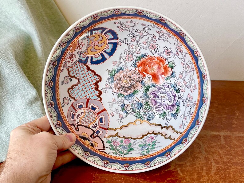 May include: A large white ceramic bowl with a blue and gold rim. The bowl is decorated with a floral pattern in shades of pink, red, blue, and green. The flowers are stylized and have a traditional Japanese look.