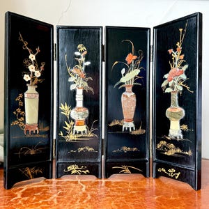 May include: A four-panel black folding screen with floral vase designs. Each panel features a different vase with colorful flowers and foliage. The vases are set against a black background with gold accents. The screen is displayed on a red-orange surface.