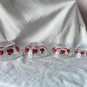 May include: Four clear glass bowls of varying sizes, each adorned with a repeating pattern of red apples with green leaves. The bowls are arranged in a nested configuration, showcasing their graduated sizes.