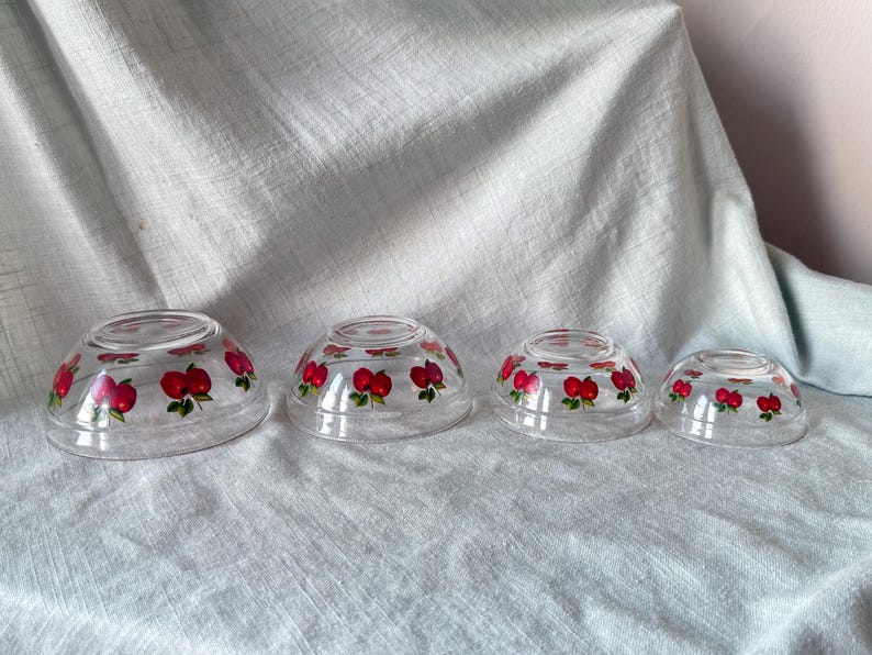 May include: Four clear glass bowls of varying sizes, each adorned with a repeating pattern of red apples with green leaves. The bowls are arranged in a nested configuration, showcasing their graduated sizes.