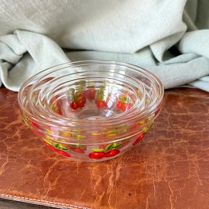 May include: A set of three clear glass bowls nested together. Each bowl has a red cherry pattern around the rim. The bowls are on a brown surface with a light green cloth in the background. Ideal for serving snacks or desserts.