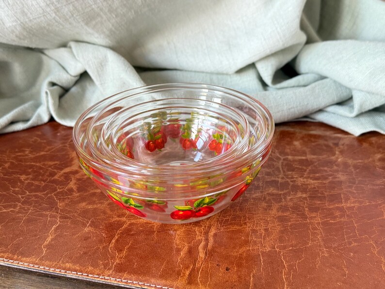 May include: A set of three clear glass bowls nested together. Each bowl has a red cherry pattern around the rim. The bowls are on a brown surface with a light green cloth in the background. Ideal for serving snacks or desserts.