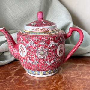 May include: A red ceramic teapot with a lid, handle, and spout. The teapot is decorated with white floral patterns and Chinese characters. The lid and handle are a darker shade of red. The teapot sits on a brown surface.