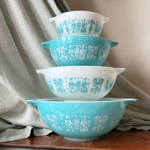 May include: A set of four stacked mixing bowls with handles, alternating between white and turquoise blue. Each bowl features a repeating white or turquoise graphic print of figures, pineapples, and farm-related imagery around the exterior.