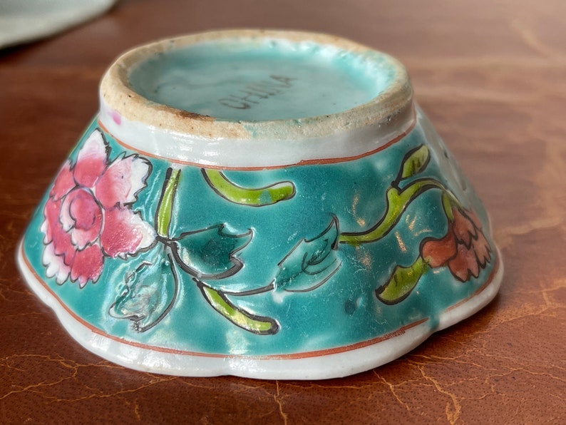 Puede incluir: Un peque&ntilde;o cuenco de porcelana con bordes festoneados, con un esmalte turquesa y un dise&ntilde;o floral. El cuenco tiene un borde blanco y las palabras "CHINA" est&aacute;n impresas en la parte inferior.