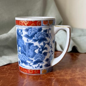 May include: A white ceramic mug featuring a blue and white floral pattern with a mythical creature, accented by red and gold geometric bands at the top and bottom rims. The handle is white with a thin gold trim along the outer edge.