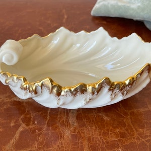 May include: A white porcelain dish shaped like a seashell with a gold rim. The dish has a wavy design and a curled handle.
