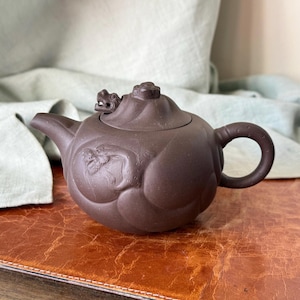 May include: Brown ceramic teapot with a dragon design. The teapot has a curved spout, a rounded handle, and a lid with a dragon head detail. The body of the teapot features an embossed dragon motif.
