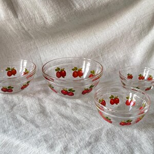 May include: Four clear glass bowls with a red apple design. The bowls are of varying sizes and are arranged on a light-colored fabric. The apple design features red apples with green leaves.