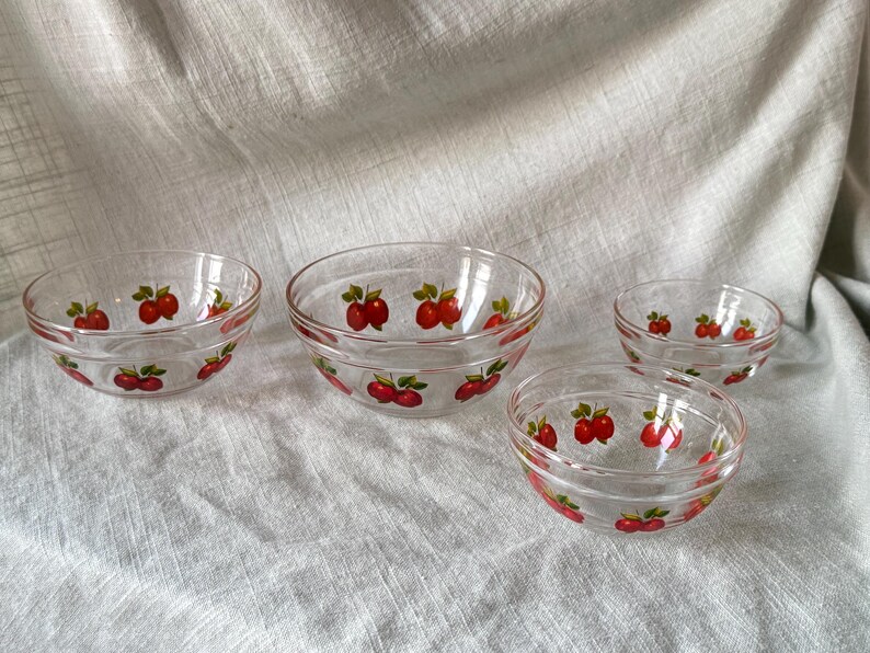 May include: Four clear glass bowls with a red apple design. The bowls are of varying sizes and are arranged on a light-colored fabric. The apple design features red apples with green leaves.