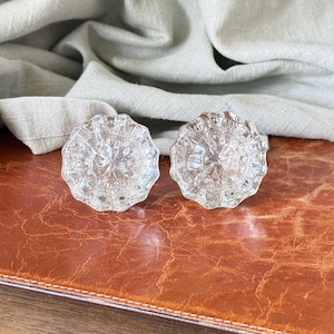 May include: Two clear glass cabinet knobs featuring a multi-faceted, starburst-like geometric pattern. Each knob has a circular shape with scalloped edges, reflecting light through the transparent material to create a bright, crystalline appearance.