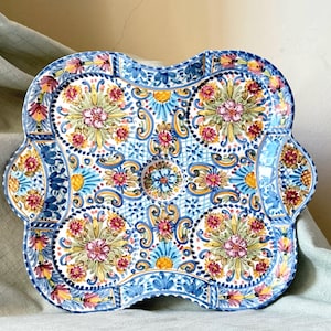 May include: A decorative, hand-painted ceramic serving tray with a scalloped edge. The tray features a floral design in shades of blue, yellow, and red. The intricate pattern includes flowers, swirls, and geometric shapes, creating a vibrant and artistic piece.