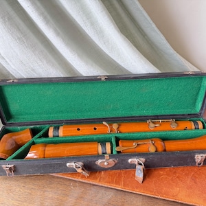 May include: A disassembled wooden flute with a polished amber finish and brass keys, resting inside a rectangular black carrying case lined with bright green felt. The case features metal latches and hinges along the front edge.