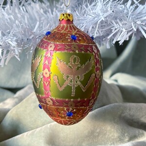 May include: An ornate Christmas ornament with a gold cap and a detailed design. The ornament features a green and gold base with pink and gold accents, and is adorned with blue jewels. It hangs from a white, tinsel-like branch.