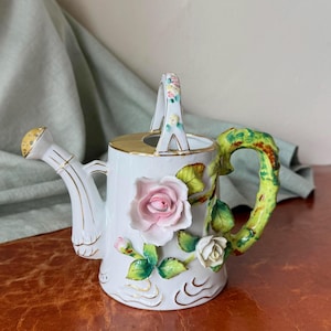 May include: White ceramic watering can with a gold rim and accents. The can features a pink rose and white rose with green leaves. The handle is green and brown, and the spout is gold. Decorative garden decor.