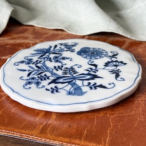 May include: A round, white ceramic coaster with a blue floral design. The coaster has a scalloped edge and is placed on a brown leather surface. The floral pattern includes flowers, leaves, and stems.