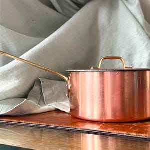 May include: A round copper saucepan with a long, slender brass handle and a matching copper lid featuring a brass loop handle. The exterior of the pan has a polished metallic finish, and the handle is securely attached with two visible rivets.