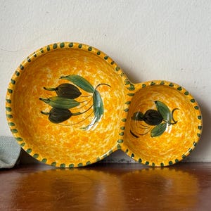 May include: A yellow ceramic serving dish with two connected bowls. The dish features a speckled pattern and is decorated with olive branch designs. The rim is adorned with green dots. Ideal for serving snacks or dips.