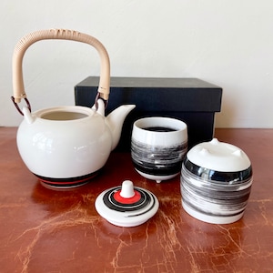May include: A white ceramic teapot with a bamboo handle and black and red accents, two white ceramic cups with black brushstroke designs, and a lid with a black and red design.