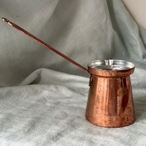May include: A copper coffee pot with a long, slender handle and a wide, flared rim. The exterior of the pot features a polished metallic copper finish with subtle variations in tone, while the interior is lined with a bright, reflective silver metal.